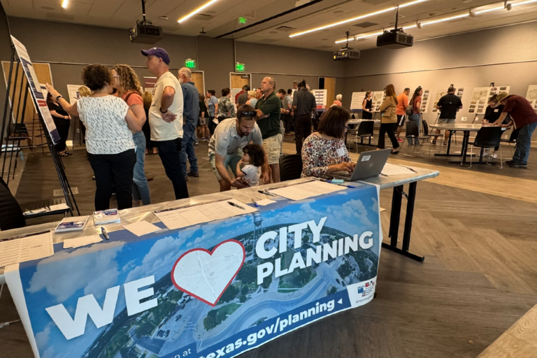 about 50 people in a room discussing display boards about zoning districts. the sign-in desk at the front has a banner reading "we heart city planning."